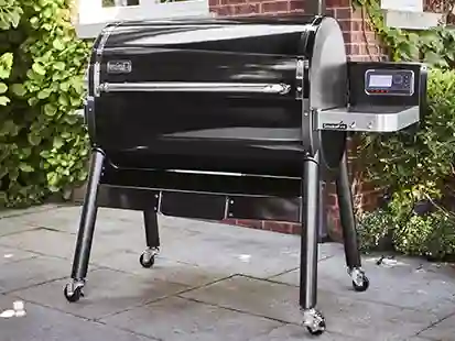 Ein schwarzer Grill steht auf Rollen auf einer Terrasse vor einer Backsteinwand und Pflanzen.